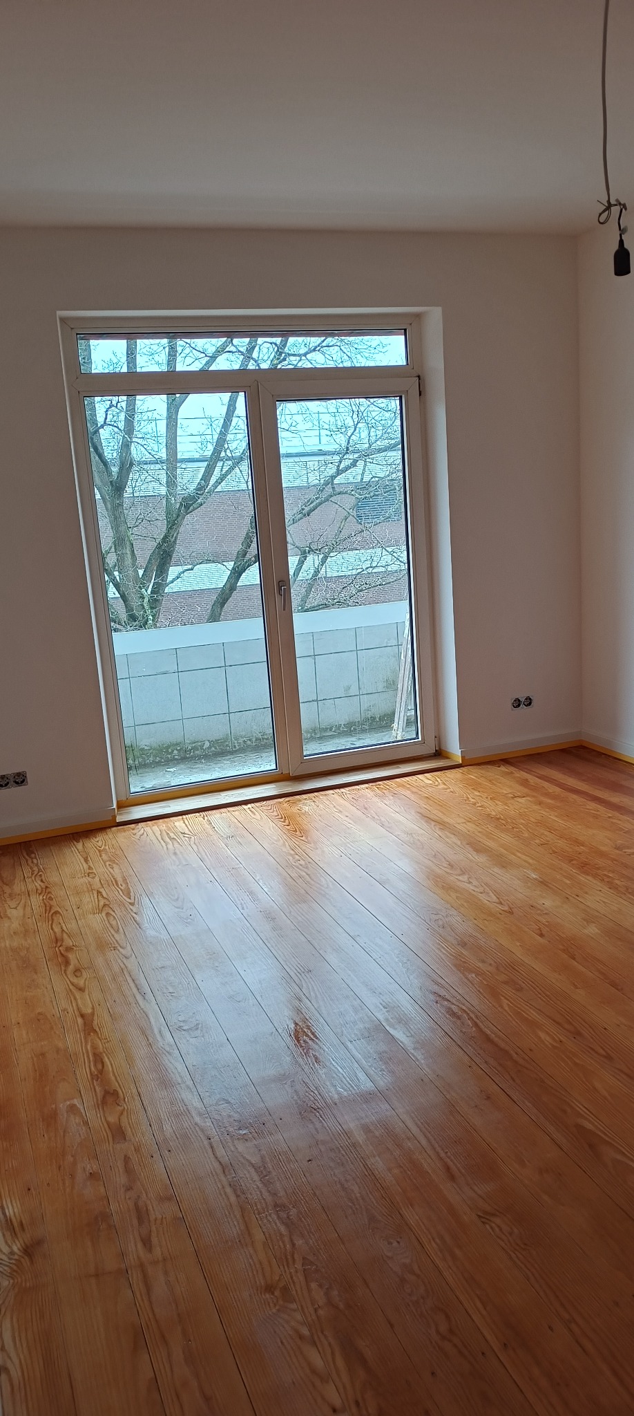 Świeżo polakierowana, błyszcząca podłoga z desek, zabezpieczona taśmą malarską przy ścianach i oknie balkonowym, widok na drzewo za oknem.