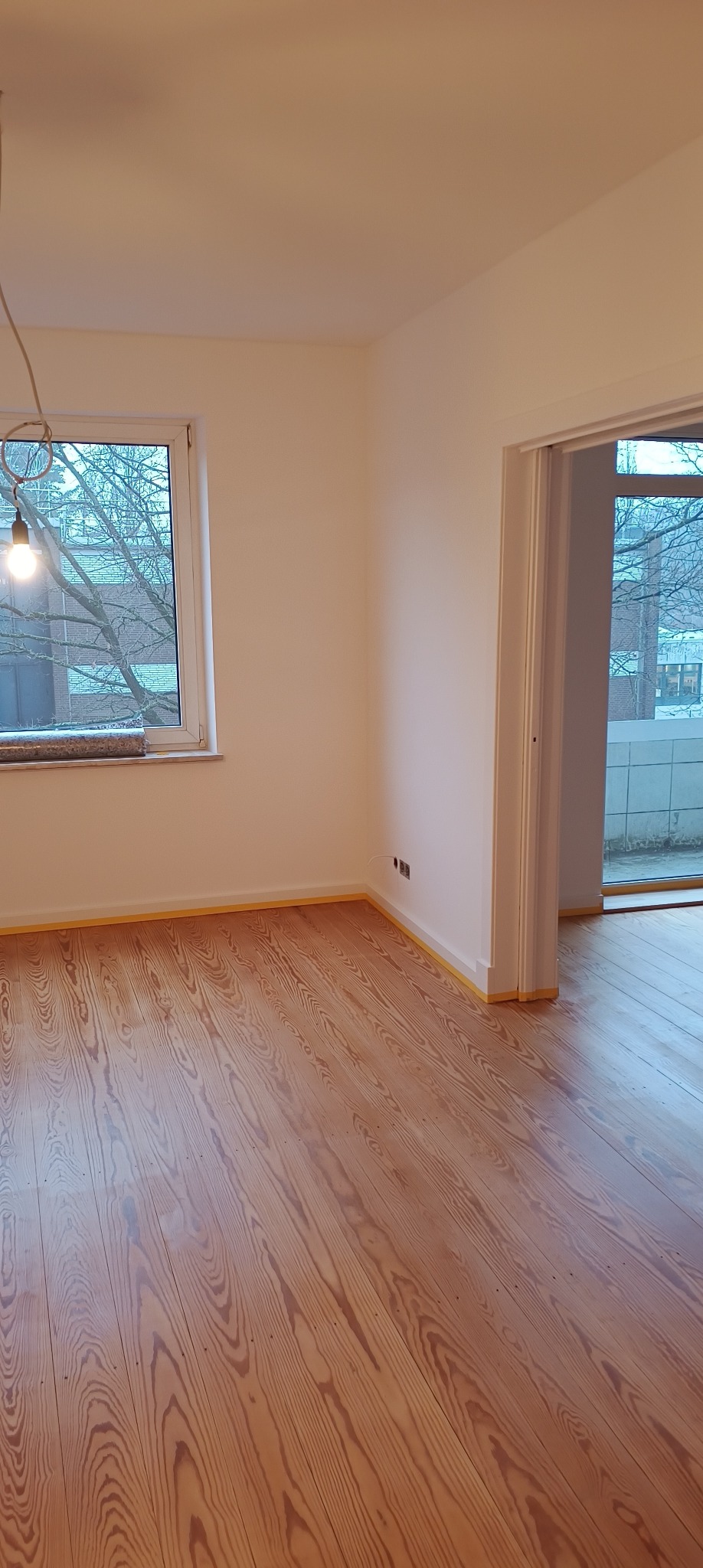 Pokój po odświeżeniu w Legnicy: świeżo pomalowane ściany na biało i odnowiona drewniana podłoga zabezpieczona taśmą malarską przy listwach.