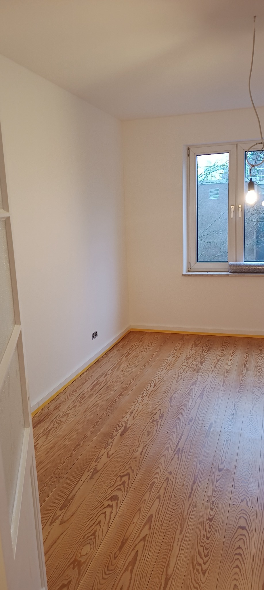 Pusty pokój z drewnianą podłogą, świeżo pomalowanymi na biało ścianami zabezpieczonymi taśmą malarską przy podłodze, widoczne gniazdka elektryczne i okno z widokiem na budynek.