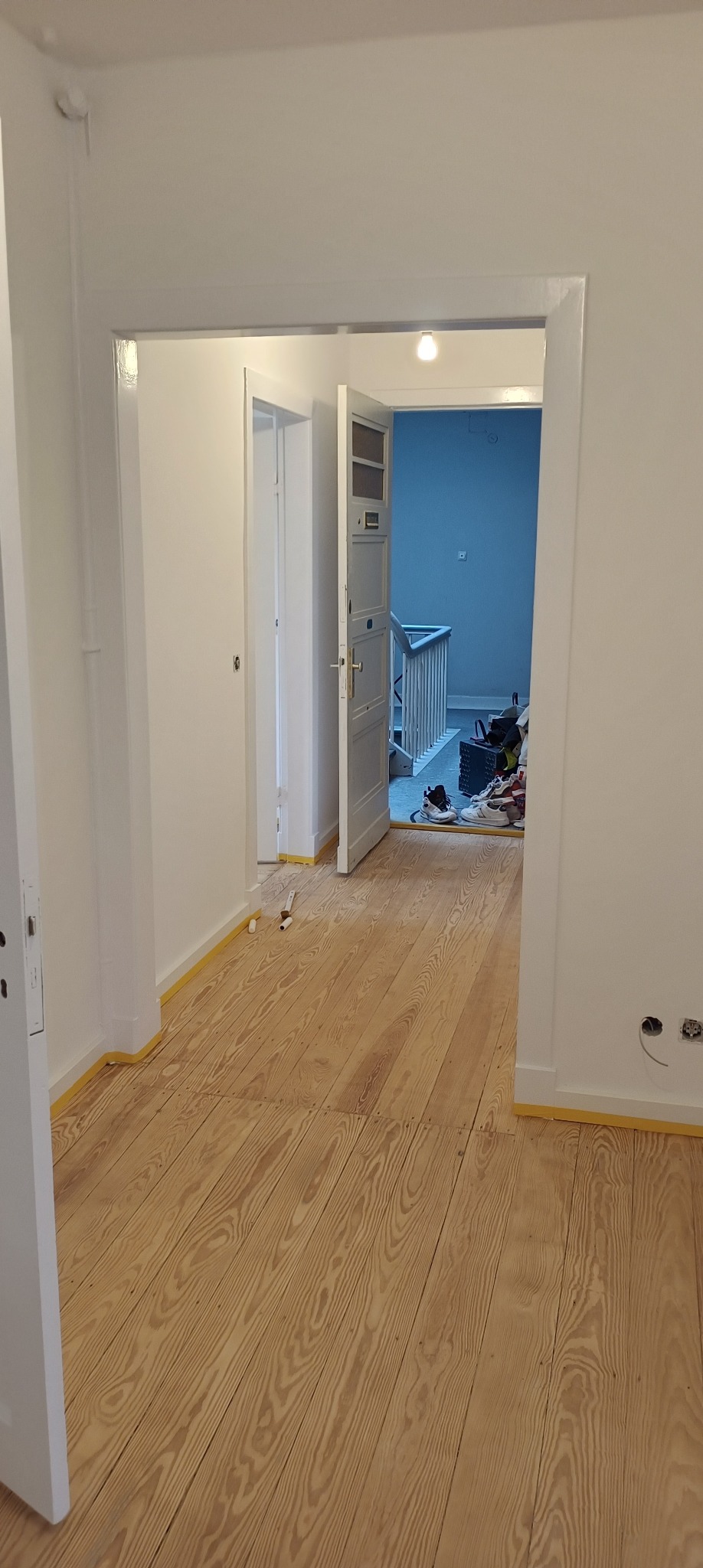 Korytarz w trakcie malowania: świeżo pomalowane białe ściany, drewniana podłoga zabezpieczona taśmą malarską, widoczne narzędzia i akcesoria malarskie w tle.