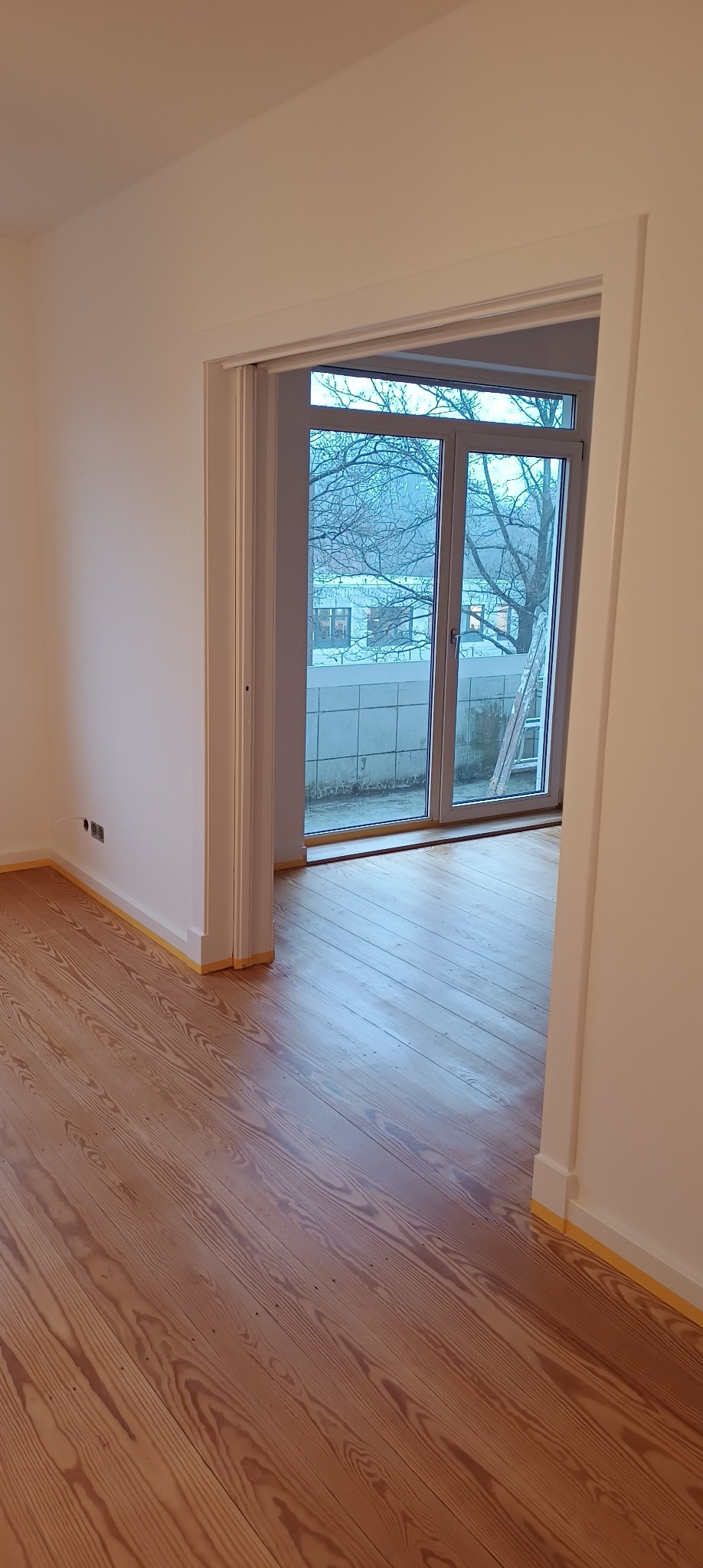 Pomalowane na biało wnętrze z drewnianą podłogą, zabezpieczoną taśmą malarską przy ścianach i framugach drzwi balkonowych; widok na balkon z drabiną opartą o ścianę budynku.