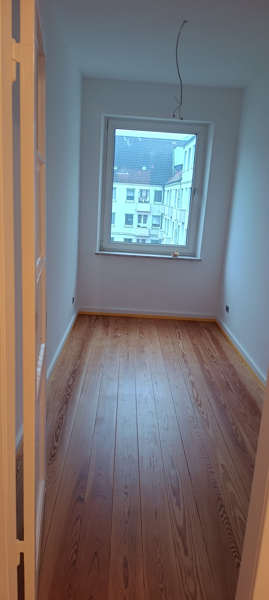 Pusty pokój z drewnianą podłogą zabezpieczoną taśmą malarską przy ścianach, świeżo pomalowane białe ściany, widok z okna na budynki mieszkalne.