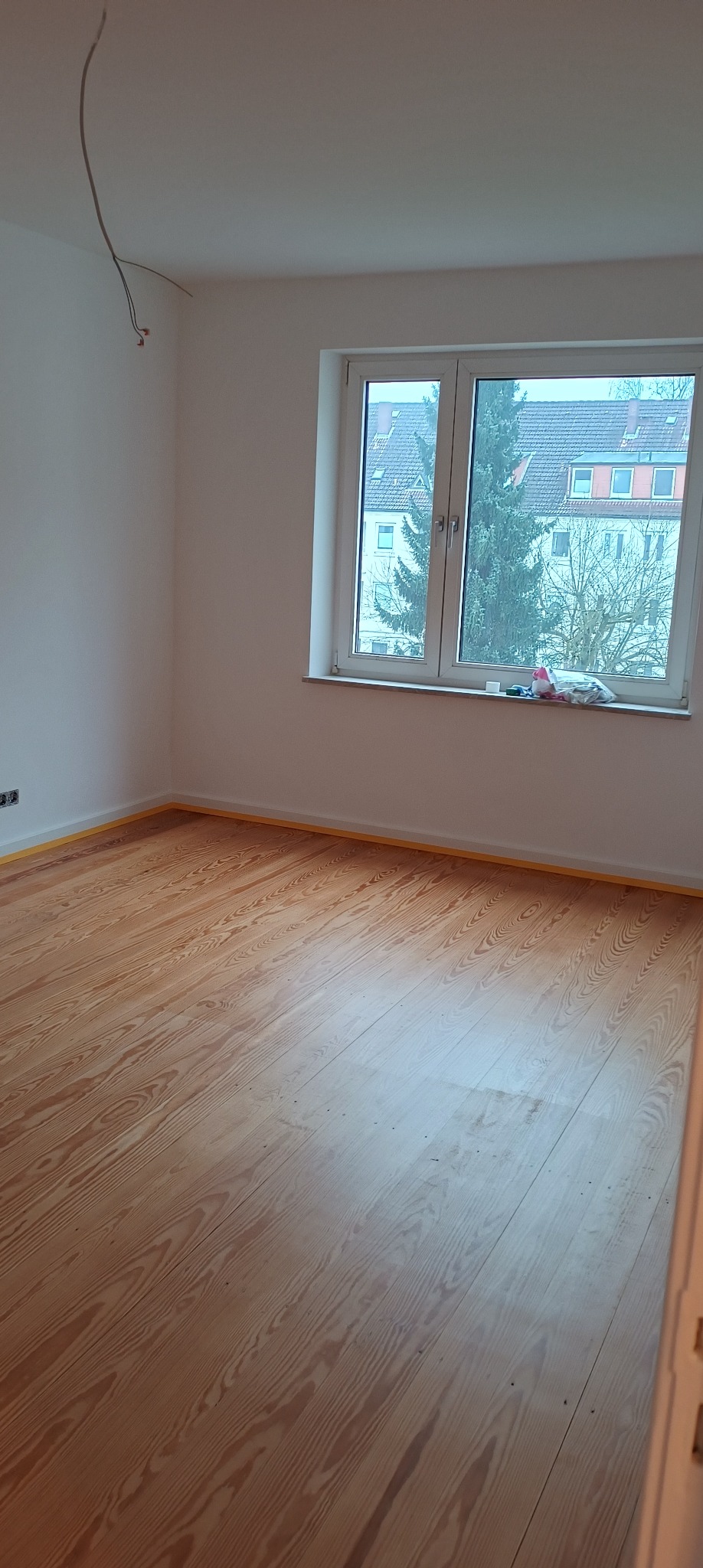 Pokój po malowaniu ścian z drewnianą podłogą zabezpieczoną taśmą malarską, widoczne niedokończone podłączenie elektryczne na suficie.