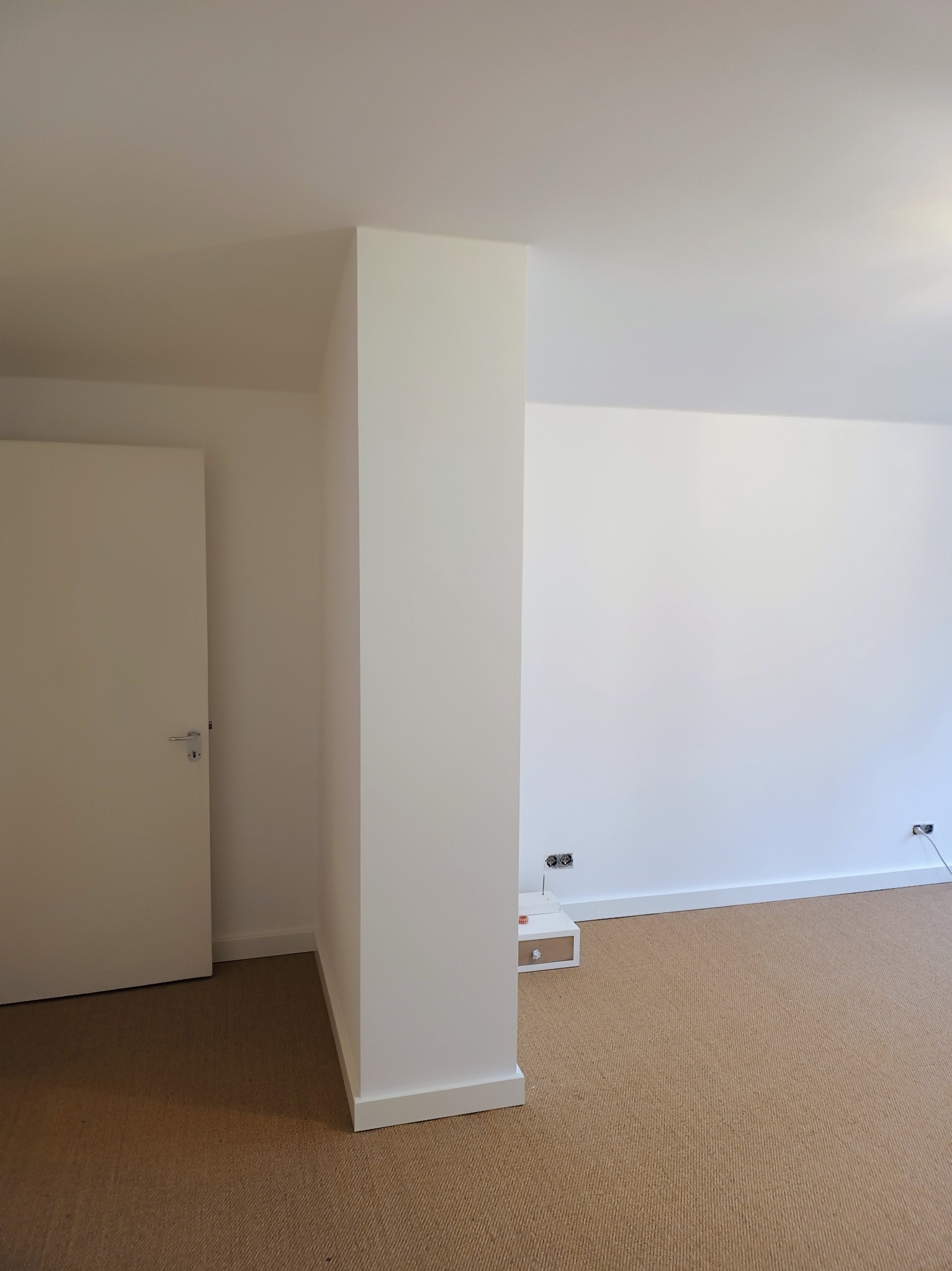 Pomalowane na biało wnętrze pokoju z beżową wykładziną, widoczne gniazdka elektryczne przy podłodze i biała szafka nocna z szufladą.