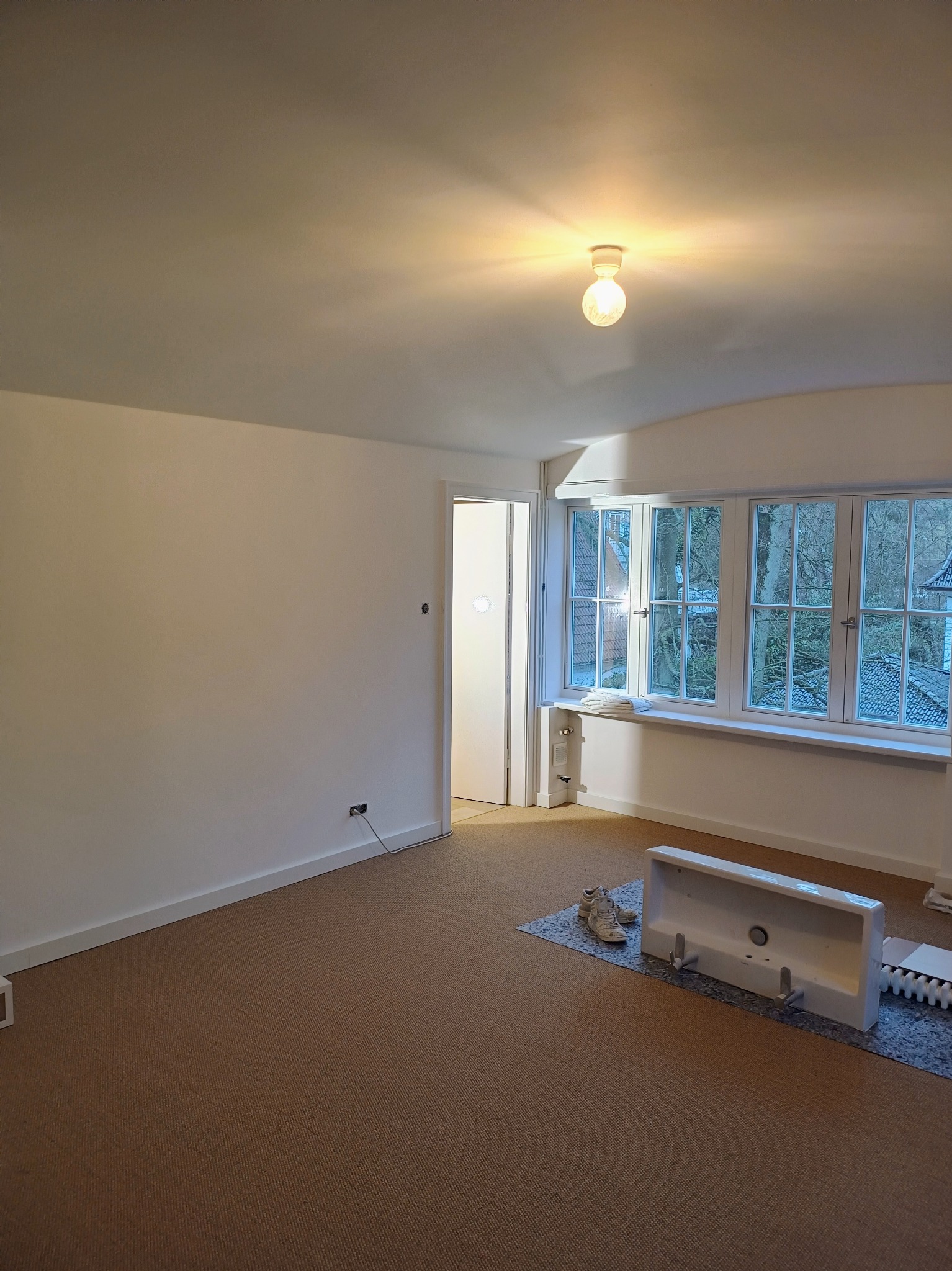 Pusty pokój z białymi ścianami, brązową wykładziną dywanową, dużym oknem i niedokończoną instalacją umywalki z grzejnikiem na podłodze.