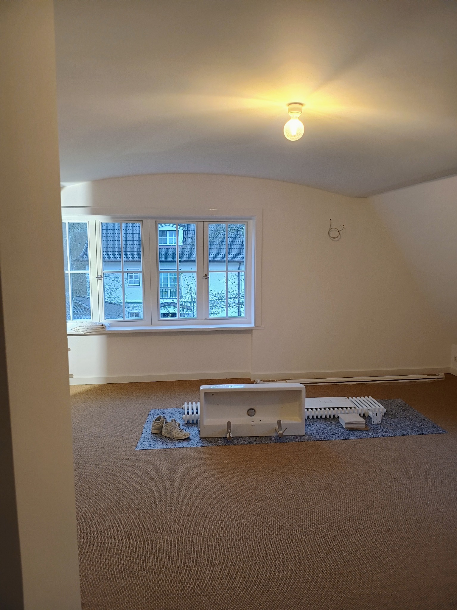 Pusta, jasna przestrzeń z oknem, na podłodze leżą odłączone elementy grzejnika i umywalka, przygotowane do montażu lub demontażu, na podłodze leżą buty.