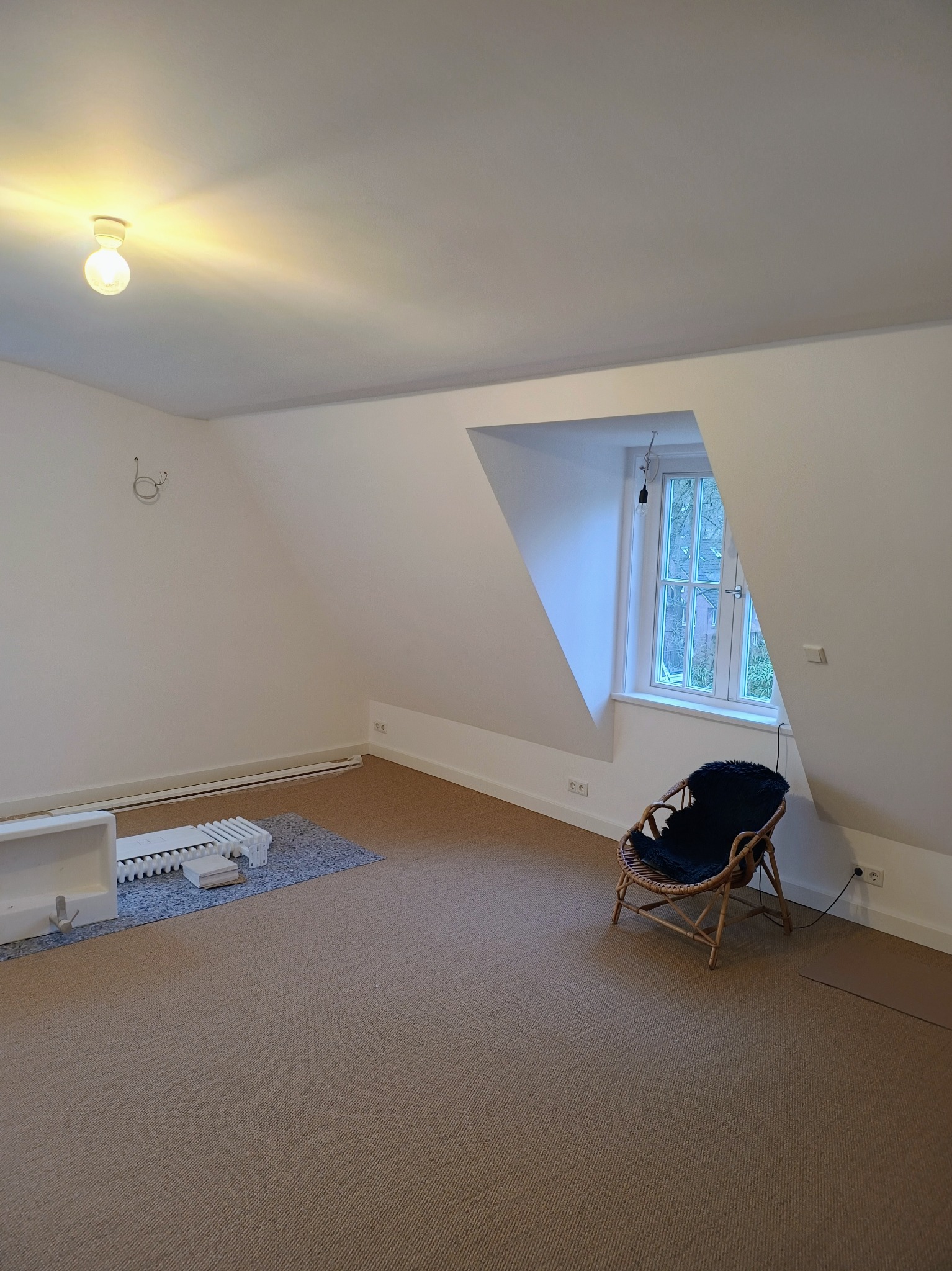 Poddasze z brązową wykładziną, wiklinowym fotelem z czarną poduszką, oknem w skośnej ścianie i grzejnikiem pod ścianą, przed rozpoczęciem prac.