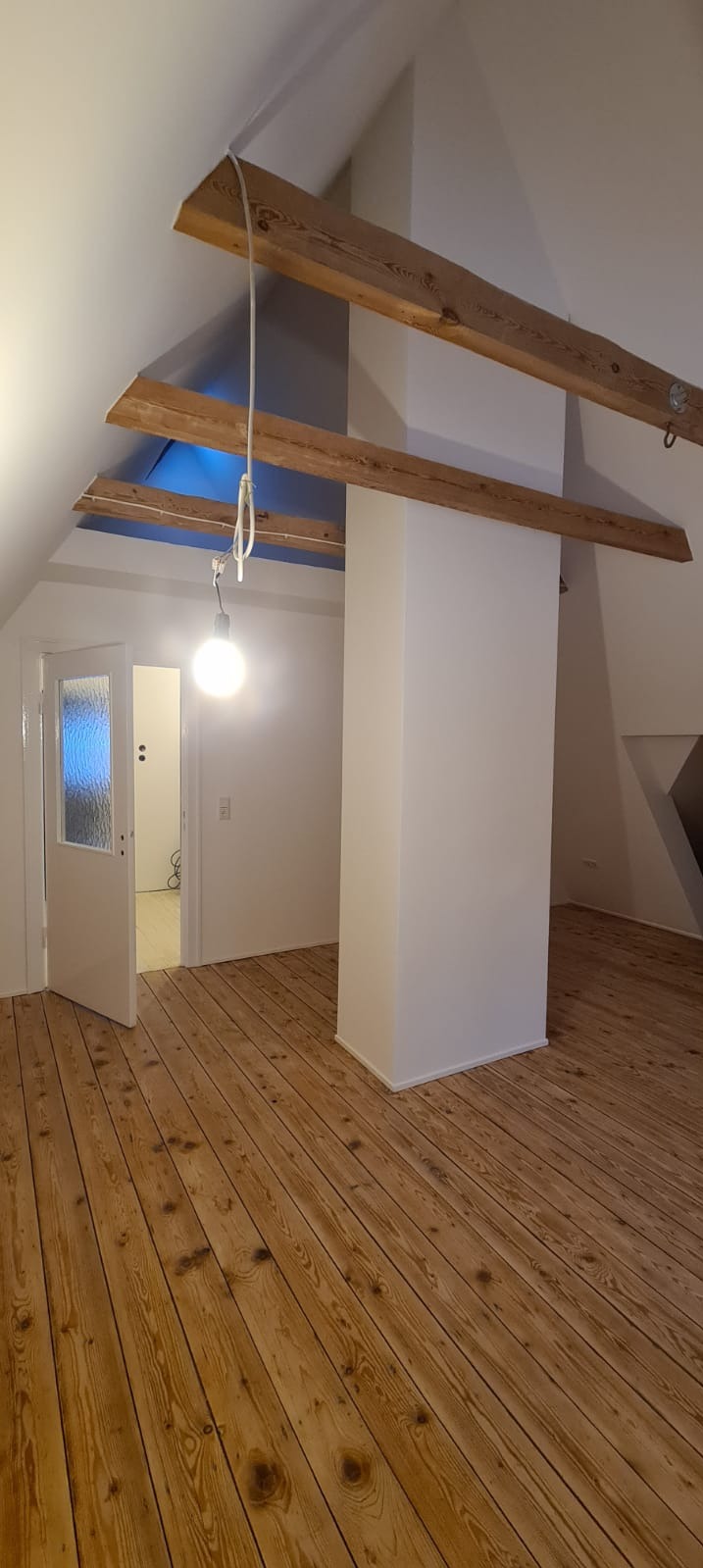 Poddasze z drewnianą podłogą, odsłoniętymi belkami stropowymi i wiszącą żarówką, widok na pomalowany filar i otwarte drzwi do kolejnego pomieszczenia.