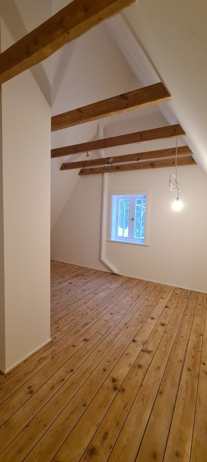 Poddasze po remoncie z drewnianą podłogą, odsłoniętymi belkami stropowymi i świeżo pomalowanymi ścianami na biało. Widoczne okno z białymi ramami i wisząca żarówka bez klosza.