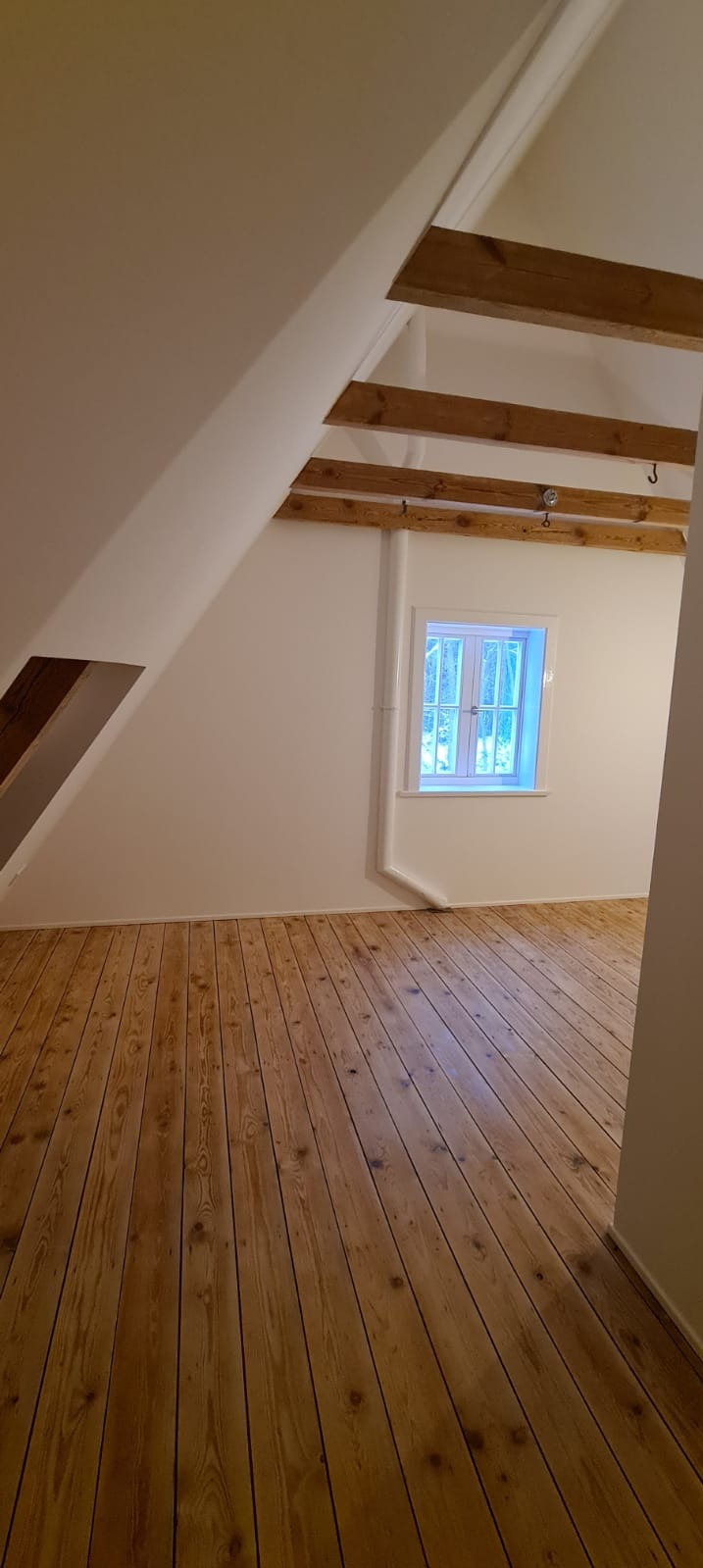 Jasne, puste poddasze z drewnianą podłogą, białymi ścianami i widocznymi belkami stropowymi, małe okno z widokiem na zieleń na zewnątrz.