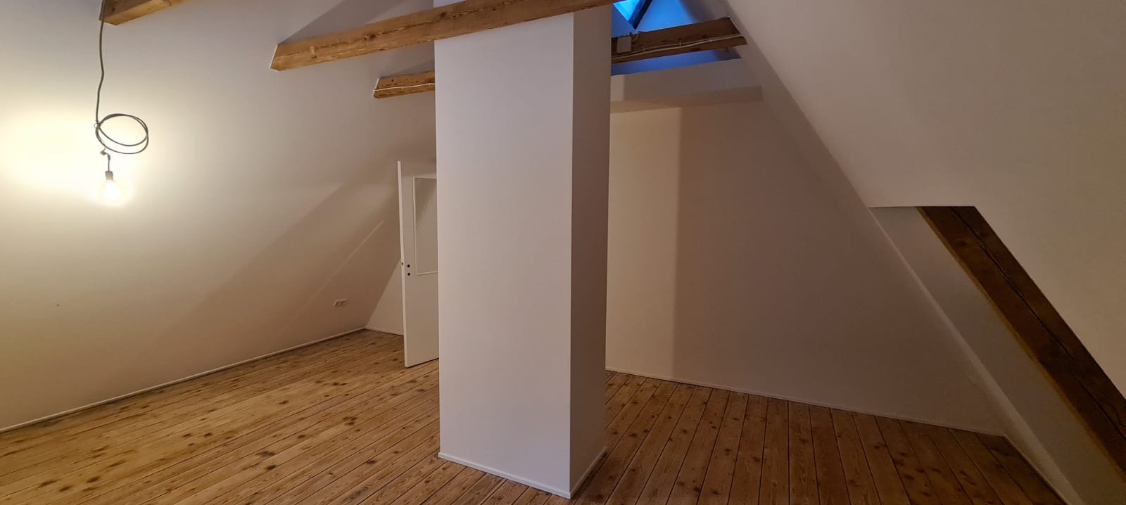 Poddasze po remoncie, świeżo pomalowane białe ściany, drewniana podłoga, widoczne drewniane belki stropowe, surowa żarówka na kablu.