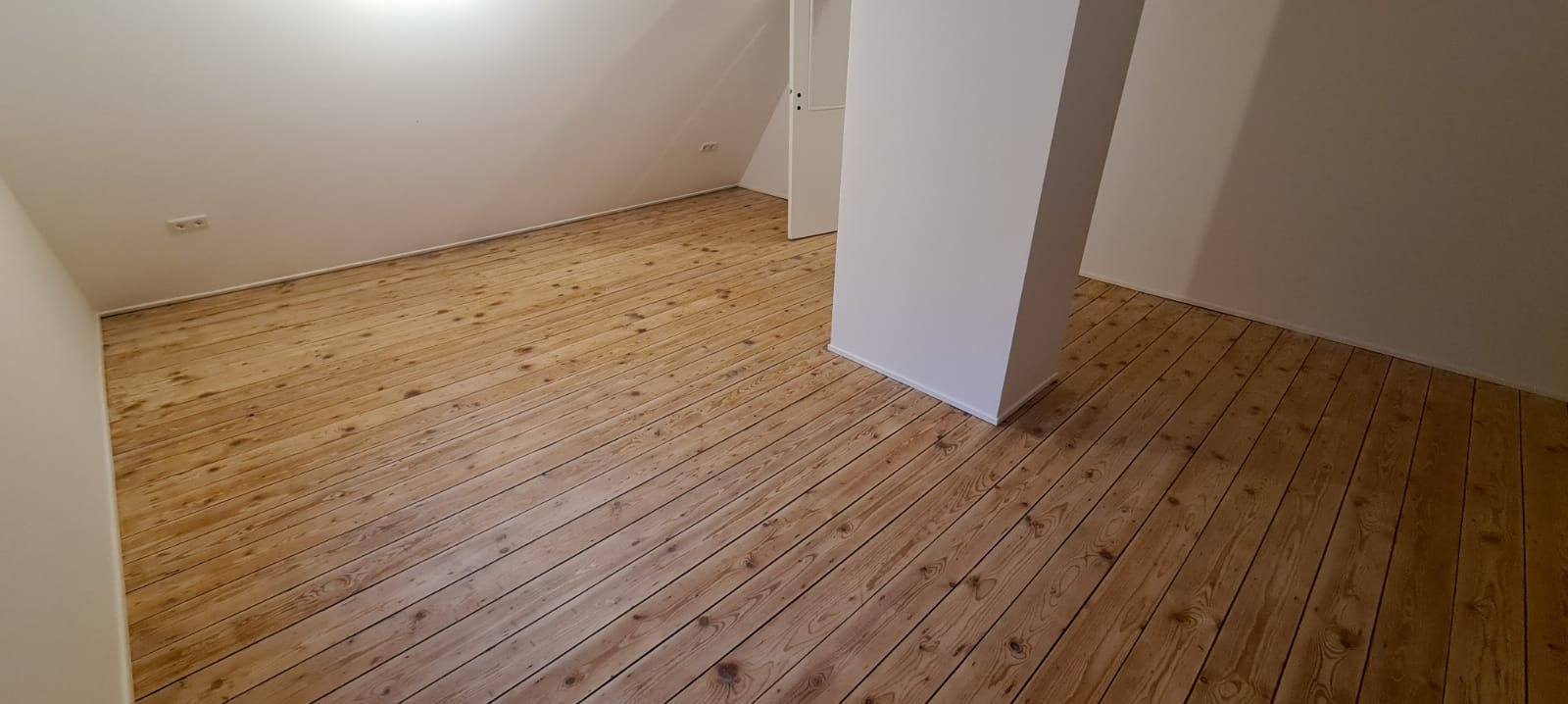 Poddasze z drewnianą podłogą z desek, białe ściany, widoczny słup nośny i otwarte drzwi w tle.