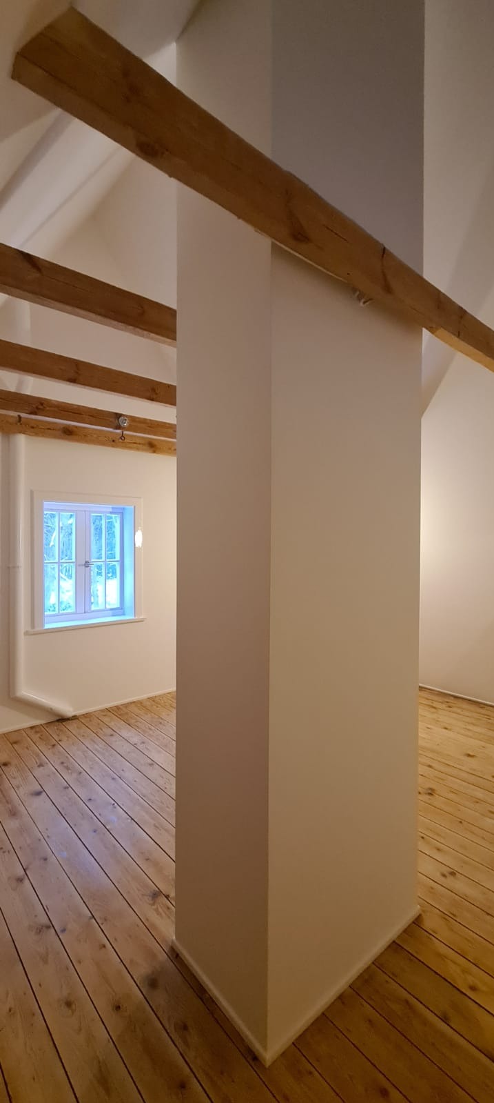 Wnętrze poddasza po remoncie: pomalowane ściany w odcieniach bieli, drewniana podłoga z widocznymi słojami, drewniane belki stropowe i okno z białymi ramami.