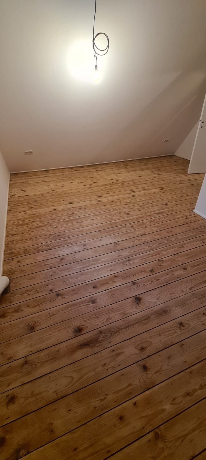 Podłoga z drewnianych paneli w jasnym odcieniu w pokoju na poddaszu, widoczne ślady użytkowania i surowy charakter drewna, prosta żarówka zwisająca z sufitu.