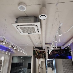 Grand Termo Norbert Moszczyński - Sufit w salonie fryzjerskim z zamontowaną kasetonową klimatyzacją, okrągłym nawiewnikiem i linią oświetleniową LED. Widoczne rury wentylacyjne i dekoracja roślinna.