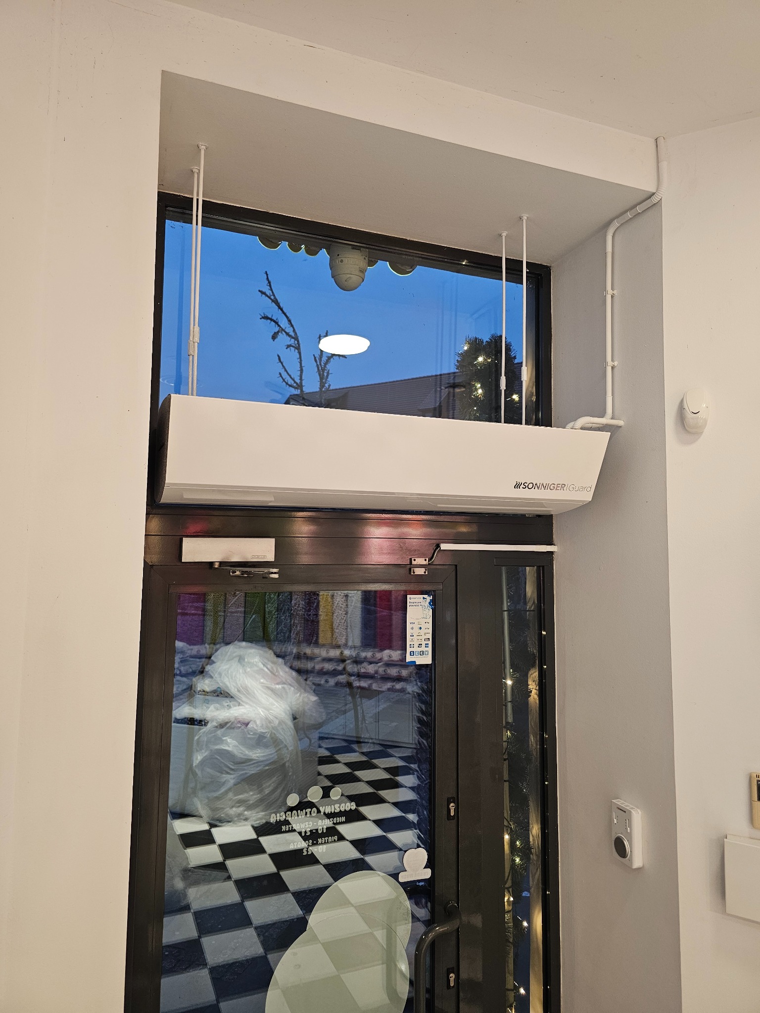 Biała kurtyna powietrzna Sonniger Guard zamontowana nad szklanymi drzwiami wejściowymi do lokalu z czarno-białą podłogą w szachownicę, widoczne przewody instalacyjne przy suficie.