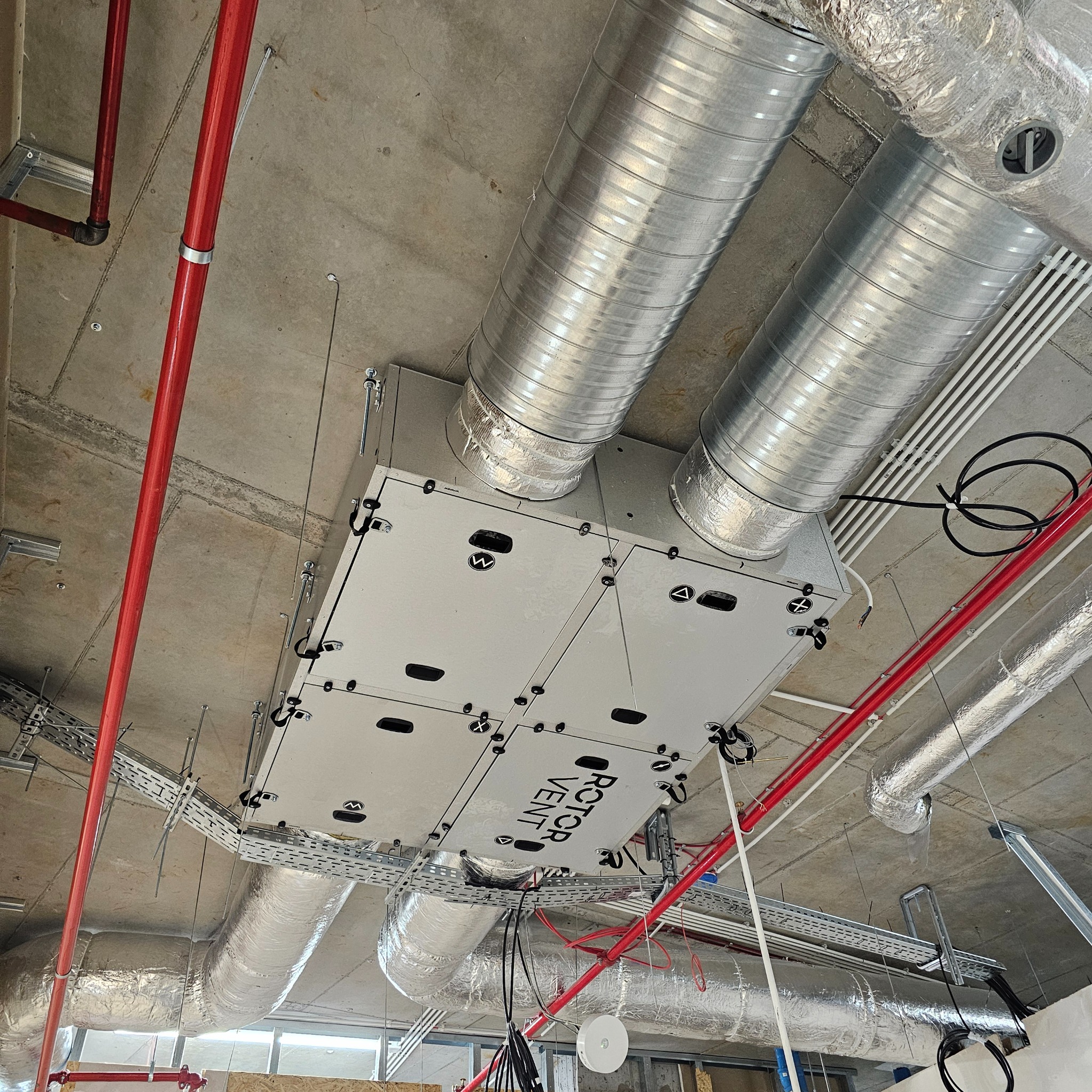 Widok z dołu na zamontowaną centralę wentylacyjną Rotor Vent z podłączonymi przewodami wentylacyjnymi i instalacjami pod sufitem w surowym stanie deweloperskim.