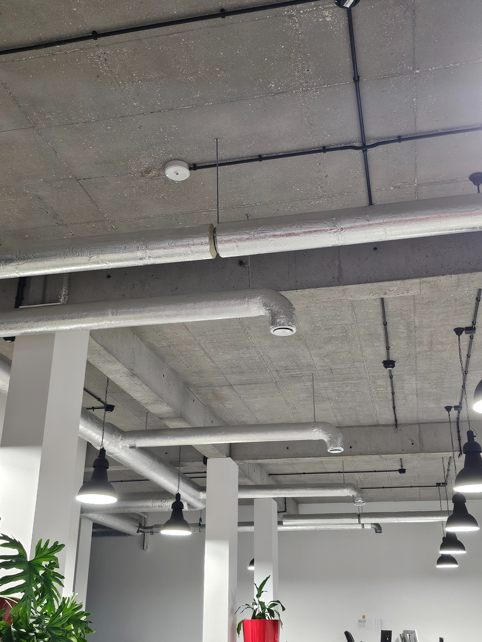 Widok na sufit biura z widocznymi instalacjami wentylacyjnymi w izolacji, czarnymi przewodami elektrycznymi oraz wiszącymi lampami przemysłowymi, betonowy sufit i białe filary