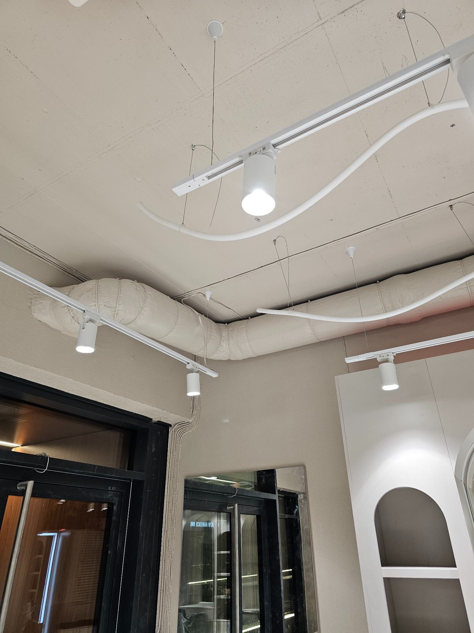 Widok na sufit z zamontowanymi szynami oświetleniowymi z reflektorami, owiniętym kanałem wentylacyjnym i instalacją elektryczną przy ościeżnicy drzwi.