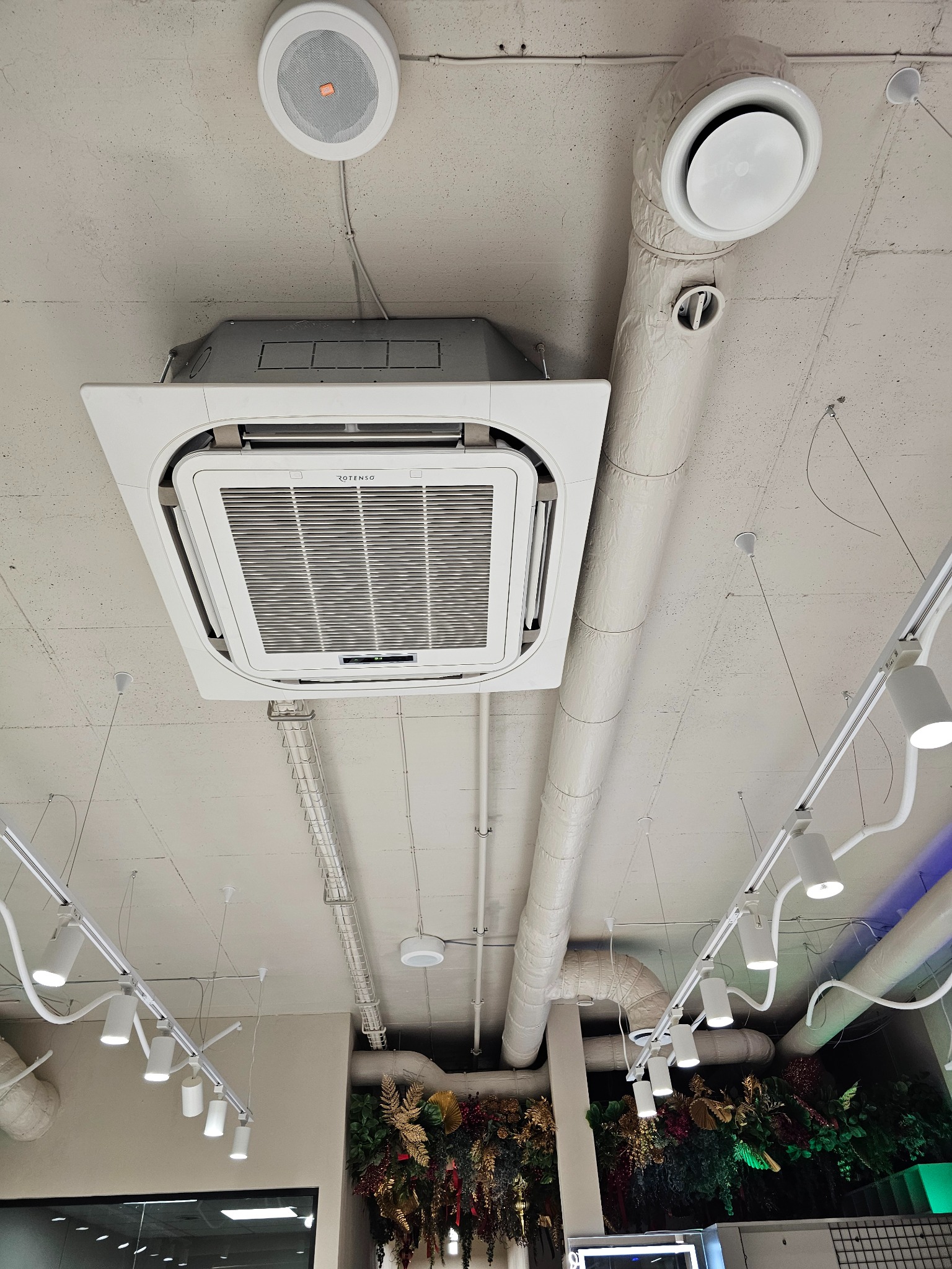 Widok z dołu na sufit biura z zamontowanym kasetonem klimatyzacyjnym marki Rotenso, okrągłym głośnikiem JBL, instalacją wentylacyjną z izolowanymi rurami, oświetleniem szynowym i elementami...