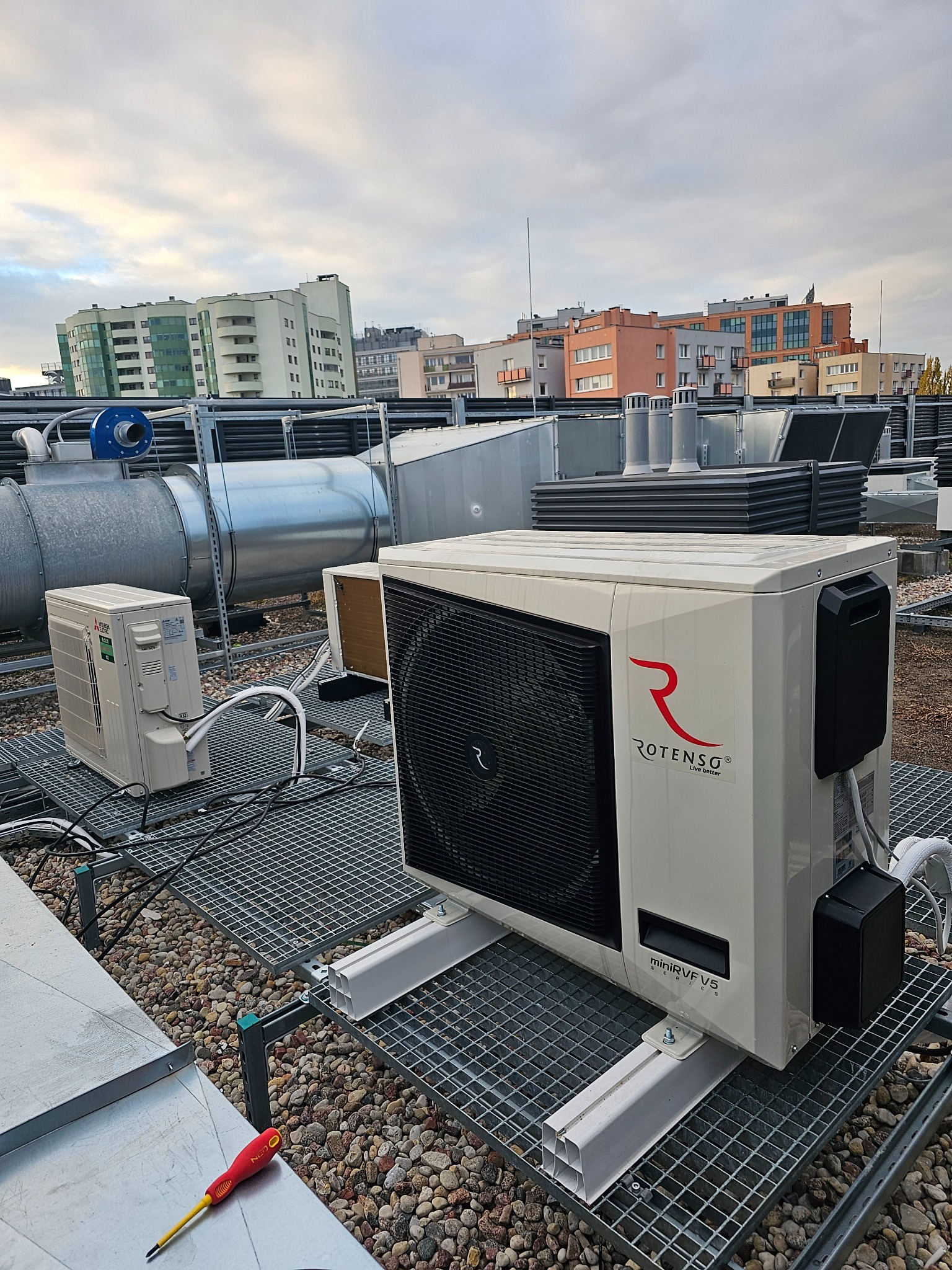 Instalacja klimatyzacji Rotenso miniRVR V5 na dachu budynku, widoczne przewody, kratownica i elementy wentylacji, w tle zabudowa miejska.