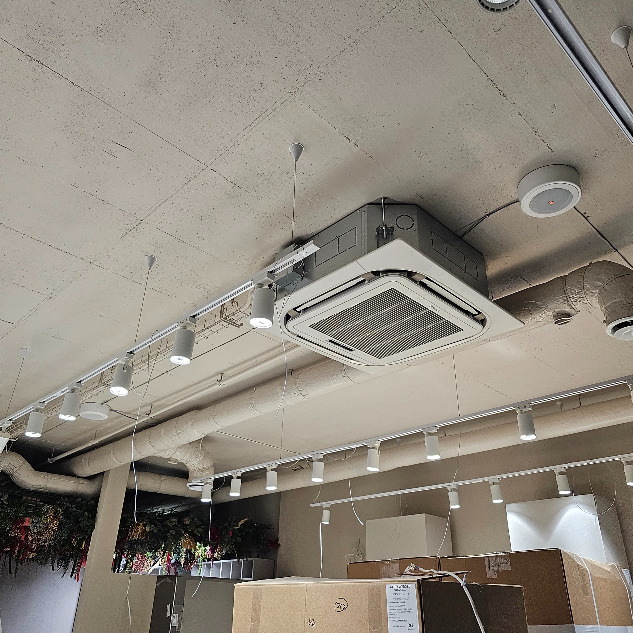 Kasetonowa jednostka klimatyzacyjna zamontowana pod sufitem w pomieszczeniu komercyjnym, widoczne oświetlenie szynowe i instalacje wentylacyjne.