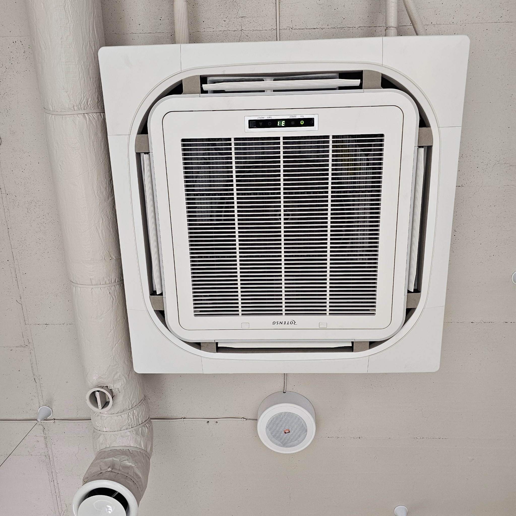 Sufitowy klimatyzator kasetonowy marki Rotenso, zamontowany na betonowym suficie, widoczne rury wentylacyjne w izolacji oraz głośnik sufitowy.