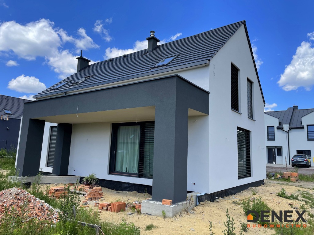 Nowoczesny dom jednorodzinny z białą elewacją i szarymi elementami architektonicznymi, widoczne cegły i teren w trakcie prac wykończeniowych.