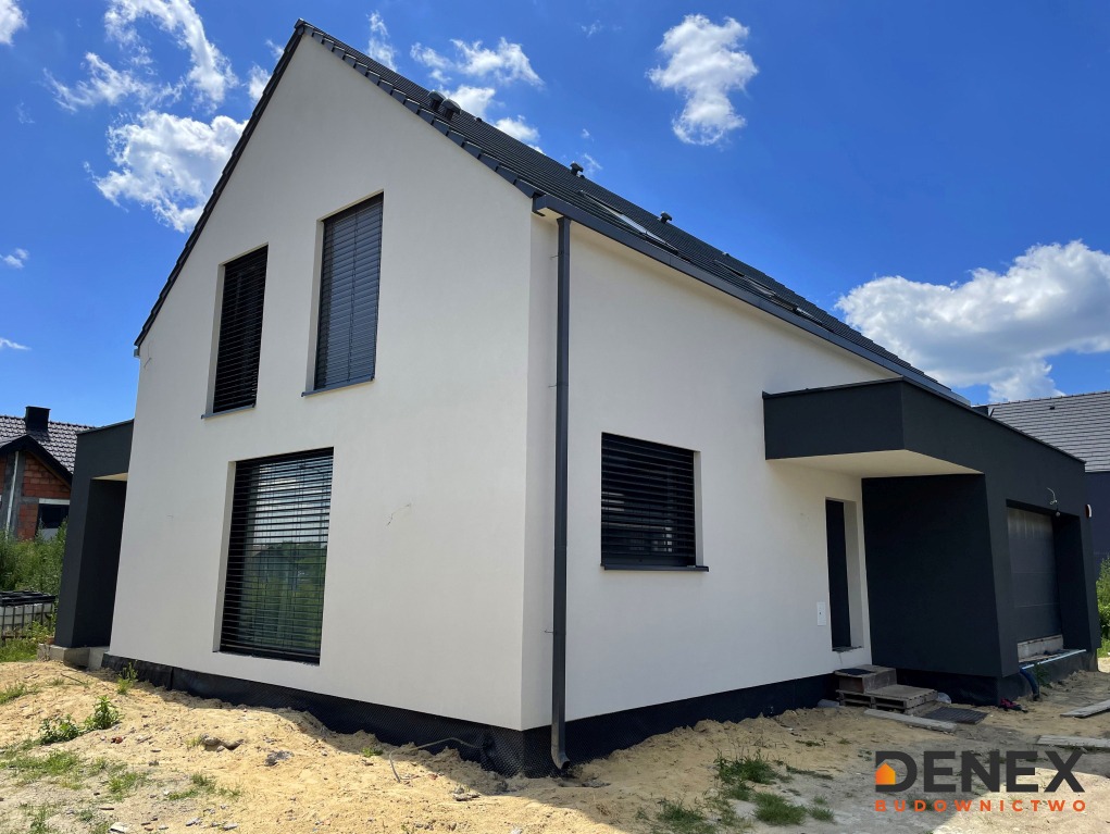 Nowoczesny dom jednorodzinny z białą elewacją i ciemnymi elementami wykończeniowymi, widok z zewnątrz w słoneczny dzień.