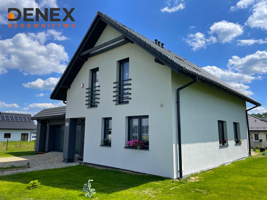 Nowoczesny dom jednorodzinny z białą elewacją, czarnymi ramami okiennymi i czarnym dachem, widok z zewnątrz na tle błękitnego nieba z chmurami, zadbany trawnik.