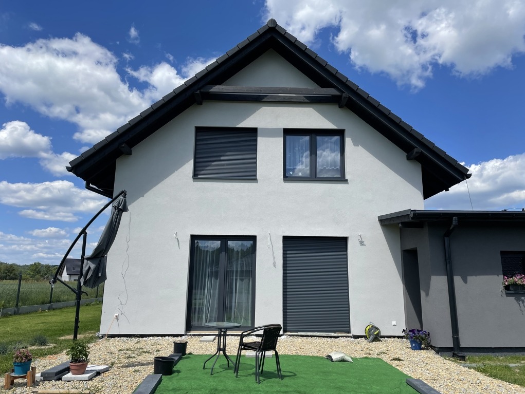 Wykończona elewacja domu jednorodzinnego z szarym tynkiem, grafitowymi oknami i roletami zewnętrznymi, widok na fragment ogrodu i błękitne niebo z chmurami.