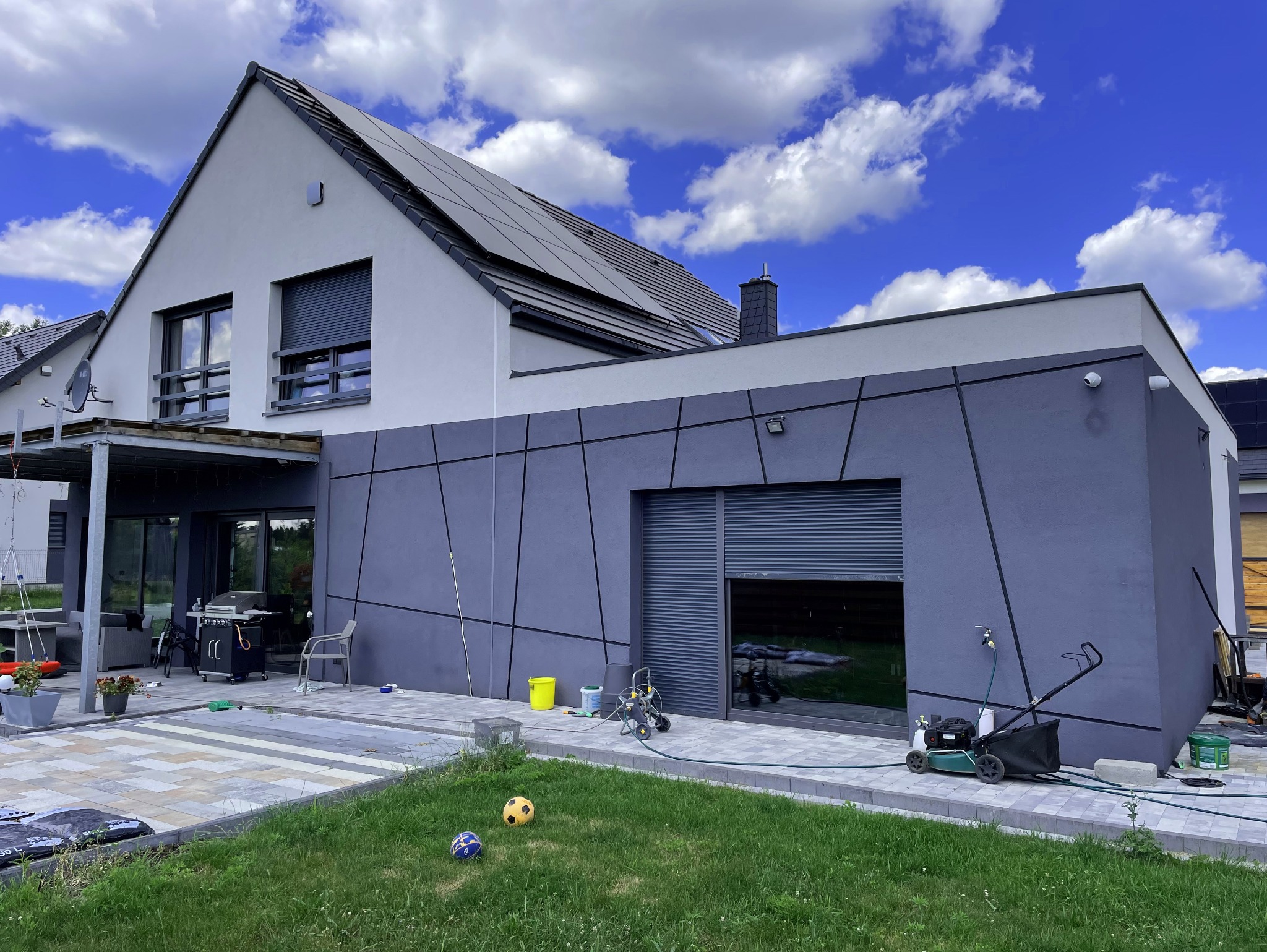 Nowoczesny dom jednorodzinny z szarą elewacją podzieloną czarnymi liniami, panele słoneczne na dachu, trawnik z piłkami i sprzęt do podlewania.