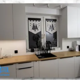 Nowoczesna, jasna kuchnia z drewnianym blatem i świątecznymi dekoracjami okiennymi. Minimalistyczny design, czarne akcenty, oświetlenie LED pod szafkami.