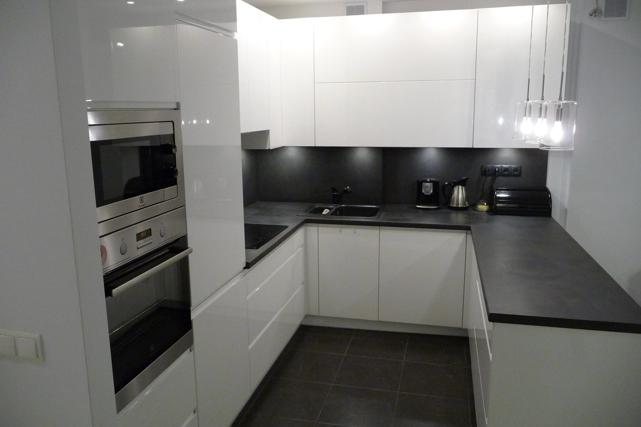 Nowoczesna, biała kuchnia z czarnym blatem i płytkami podłogowymi. Wbudowany piekarnik Electrolux. Minimalistyczne oświetlenie nad blatem. Błyszczące fronty szafek.
