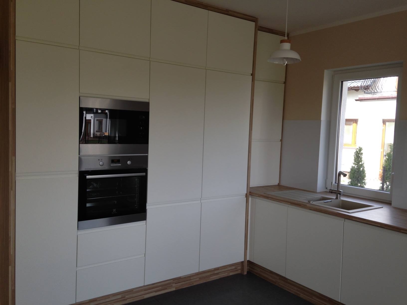 Biała zabudowa kuchenna z piekarnikiem i szafkami wiszącymi, drewniane akcenty, widok na zewnątrz przez okno, nowoczesne oświetlenie.