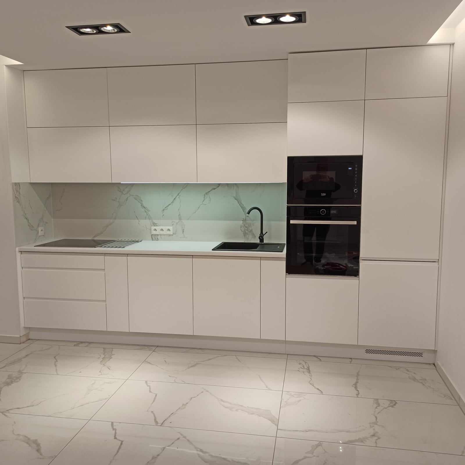 Minimalistyczna, biała kuchnia z marmurowym backsplashiem i czarną armaturą. Wbudowane sprzęty AGD, płytki podłogowe imitujące marmur. Oświetlenie sufitowe.