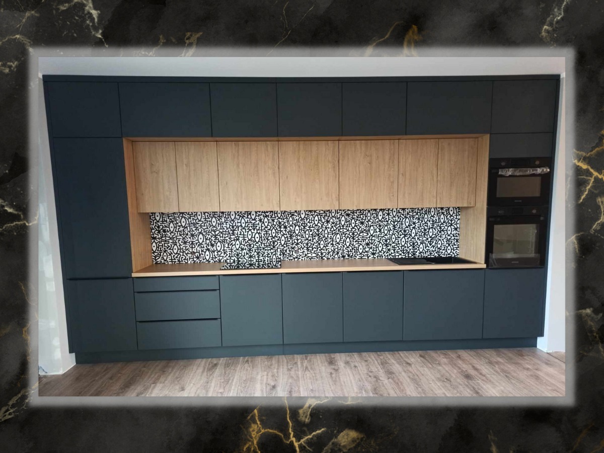 Nowoczesna kuchnia z grafitowymi szafkami, drewnianymi akcentami i wzorzystą mozaiką nad blatem. Zabudowa z piekarnikiem i mikrofalówką, jasna podłoga.