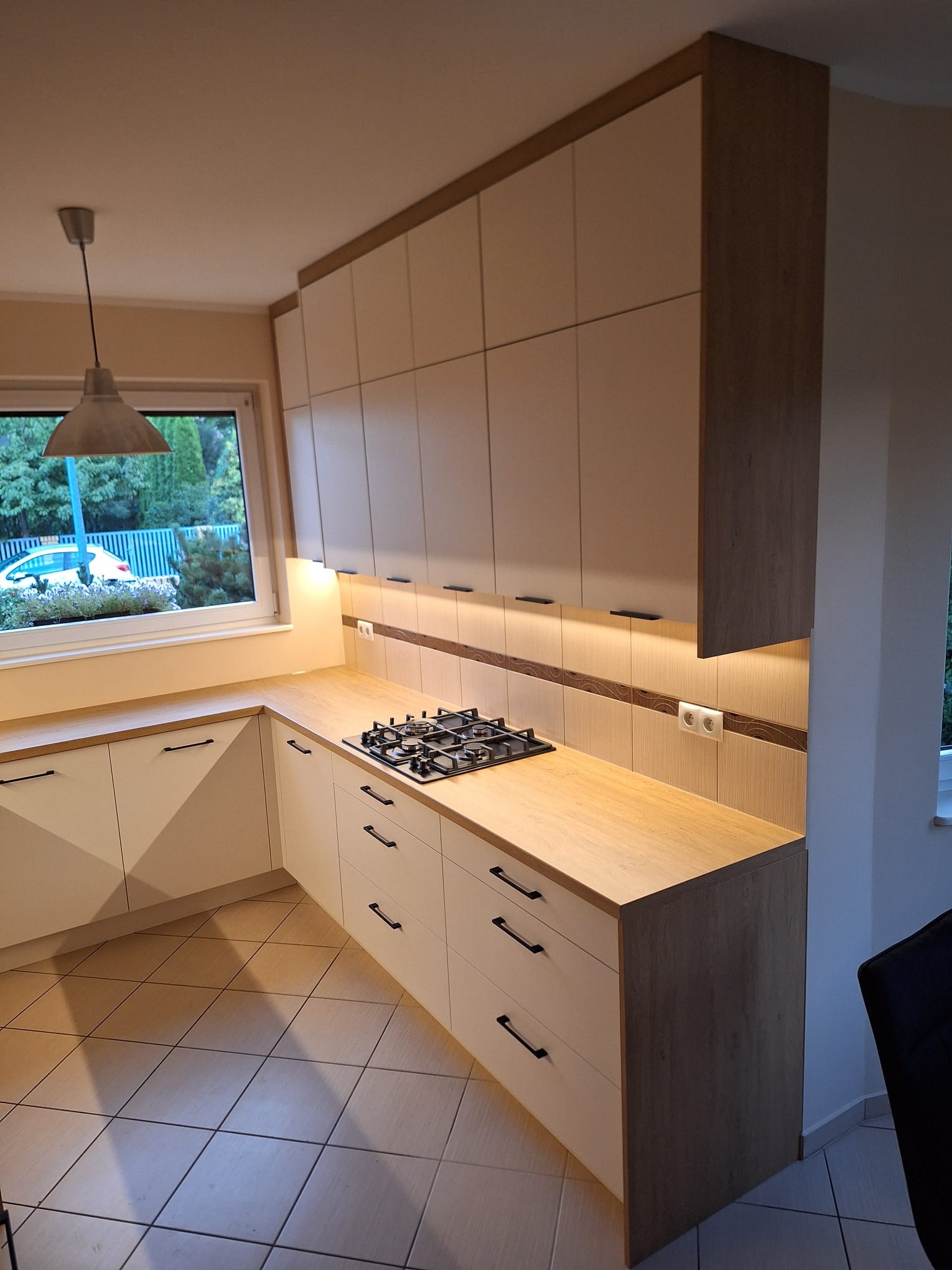 Nowoczesna kuchnia z beżowymi szafkami i drewnianym blatem, czarne uchwyty, płyta gazowa, oświetlenie LED pod szafkami, widok na okno z zielenią na zewnątrz.