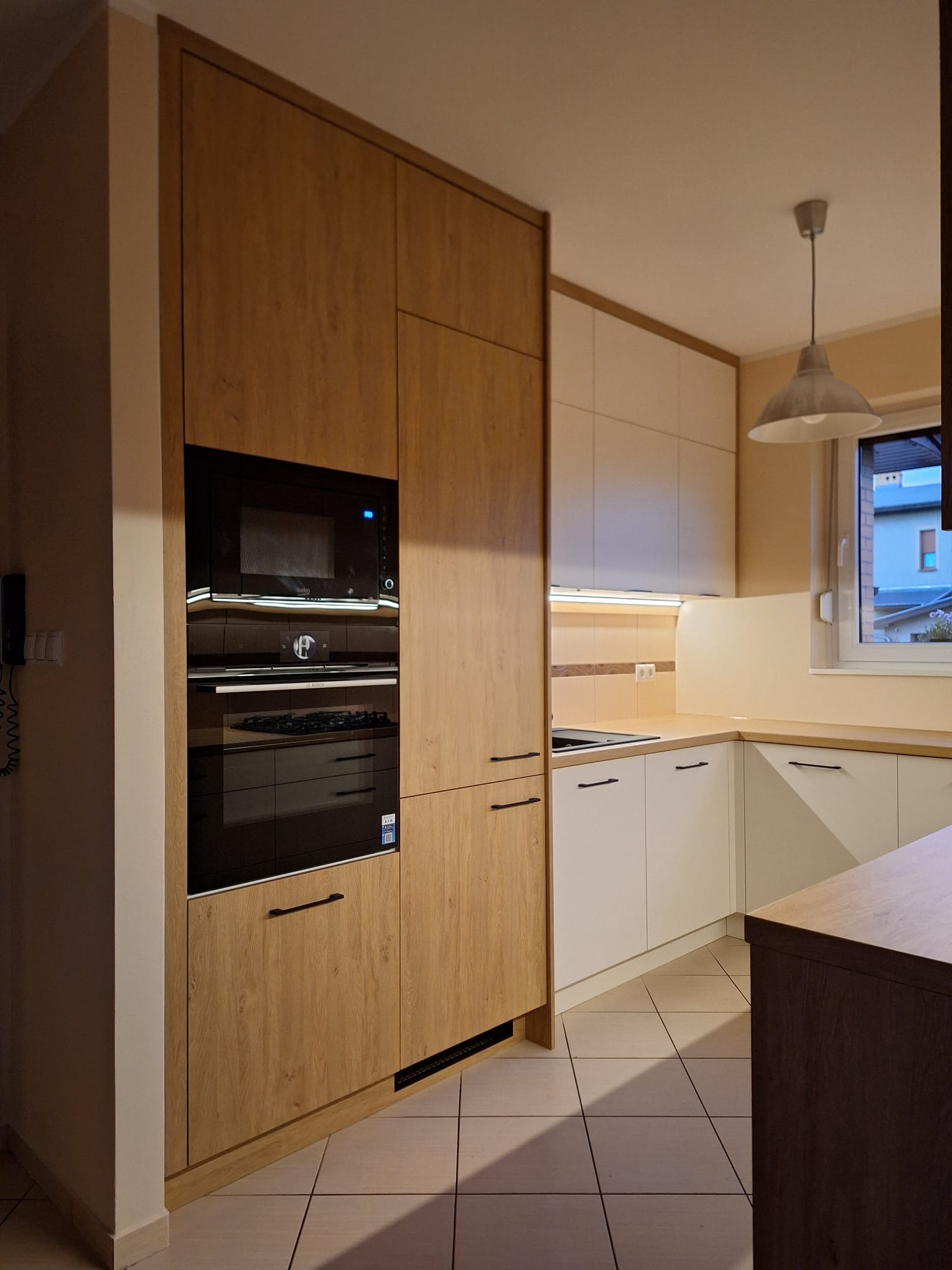 Nowoczesna kuchnia: zabudowa z drewnopodobnymi frontami, czarny piekarnik i mikrofalówka, białe szafki dolne, minimalistyczne uchwyty, płytki podłogowe.