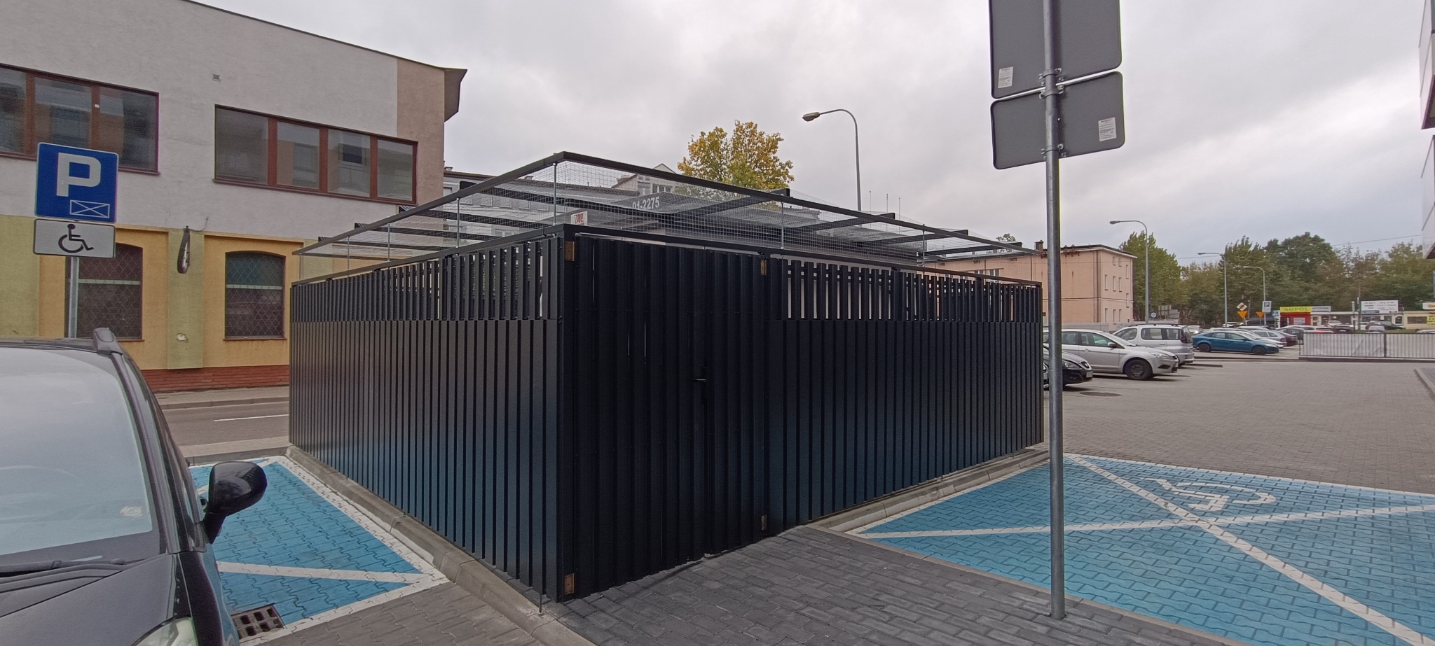 Nowoczesna, czarna, metalowa konstrukcja z ażurowym dachem na parkingu miejskim. Widoczne pionowe panele i siatka. W tle budynki i zaparkowane samochody.