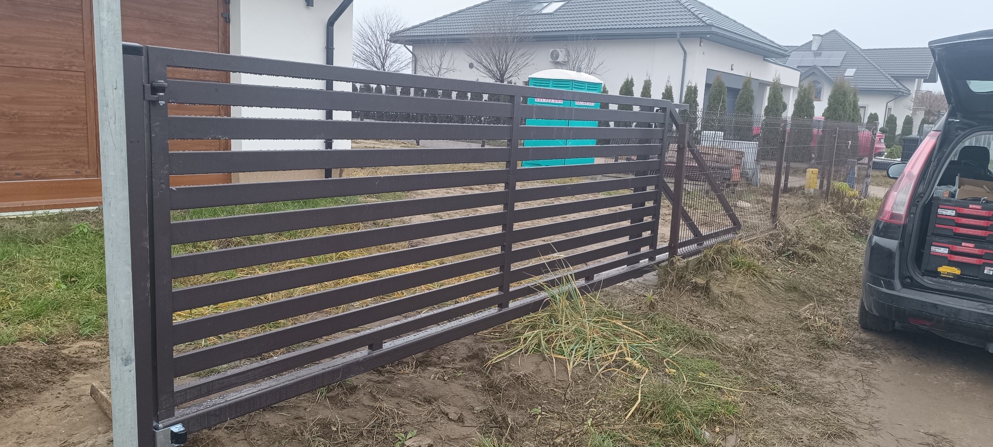 Nowoczesna, brązowa brama przesuwna z poziomymi panelami, częściowo zamontowana. W tle widoczny dom i samochód z narzędziami.