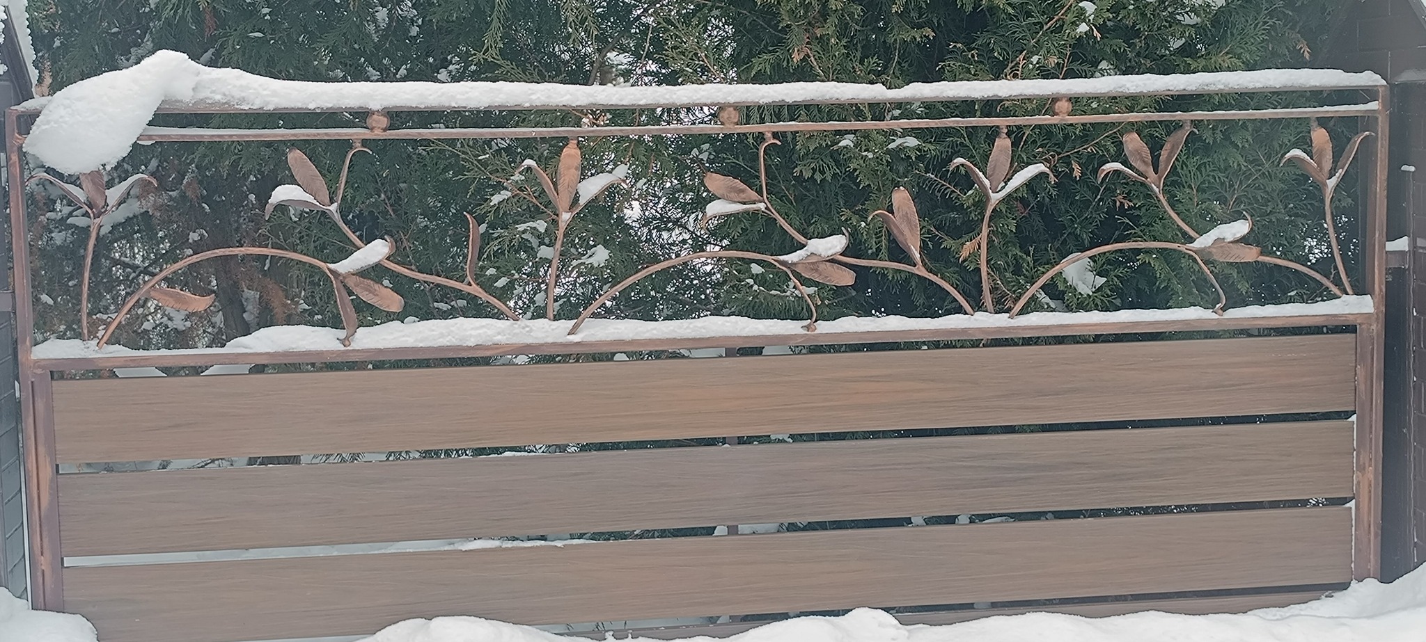 Dekoracyjne ogrodzenie z drewnopodobnymi panelami i metaloplastyką w kształcie liści, pokryte śniegiem. Tło stanowi zielony żywopłot.