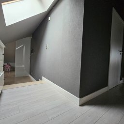 Er-Bud Eryk Skomro - Poddasze z oknem dachowym i panelami podłogowymi w odcieniach szarości; ściana pomalowana w dwóch odcieniach szarości, widoczna biała listwa przypodłogowa i białe drzwi.