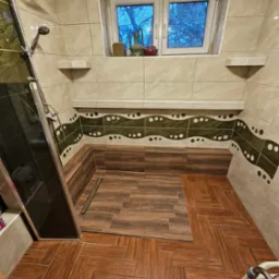 Wykończona łazienka z brązowymi płytkami imitującymi drewno na podłodze i w strefie prysznica, beżowe płytki na ścianach z dekoracyjnym pasem w odcieniach zieleni i bieli, okno z widokiem na drzewa...