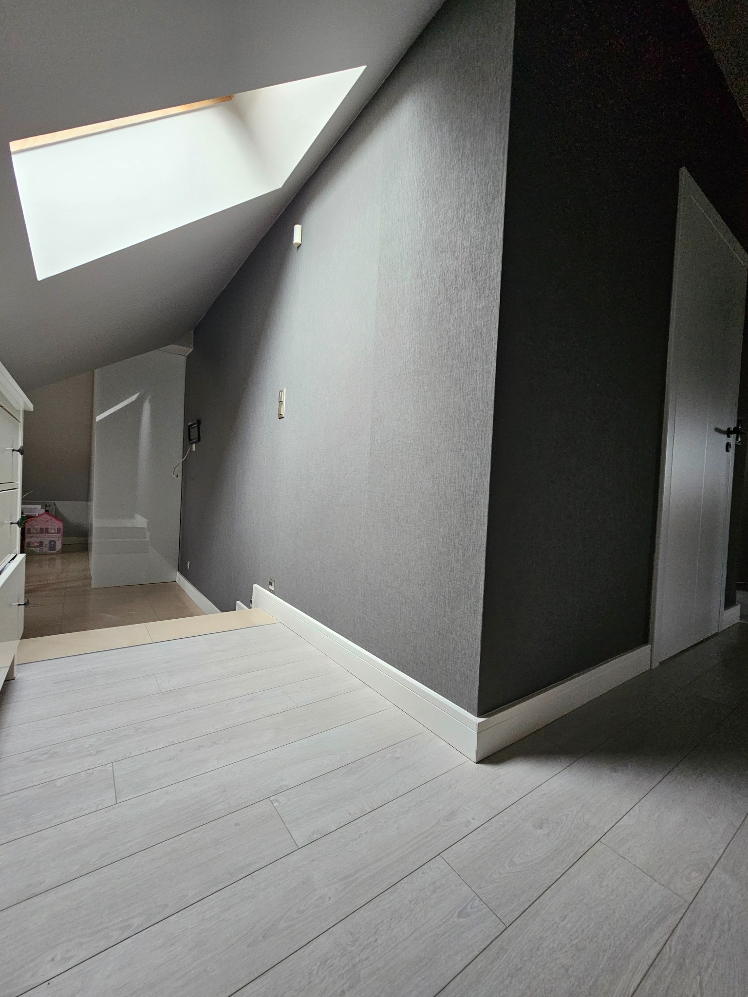 Poddasze z oknem dachowym i panelami podłogowymi w odcieniach szarości; ściana pomalowana w dwóch odcieniach szarości, widoczna biała listwa przypodłogowa i białe drzwi.