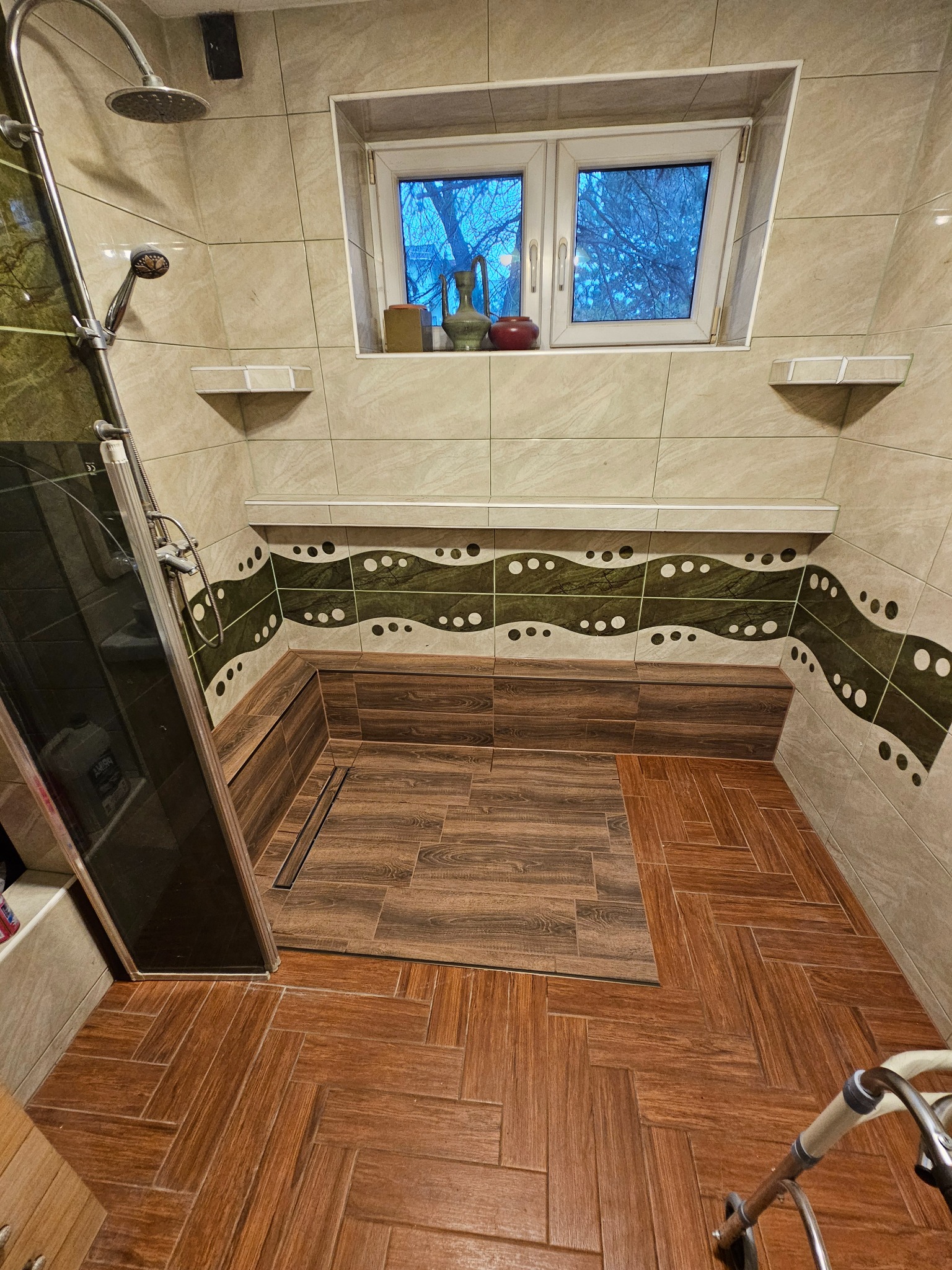 Wykończona łazienka z brązowymi płytkami imitującymi drewno na podłodze i w strefie prysznica, beżowe płytki na ścianach z dekoracyjnym pasem w odcieniach zieleni i bieli, okno z widokiem na drzewa...
