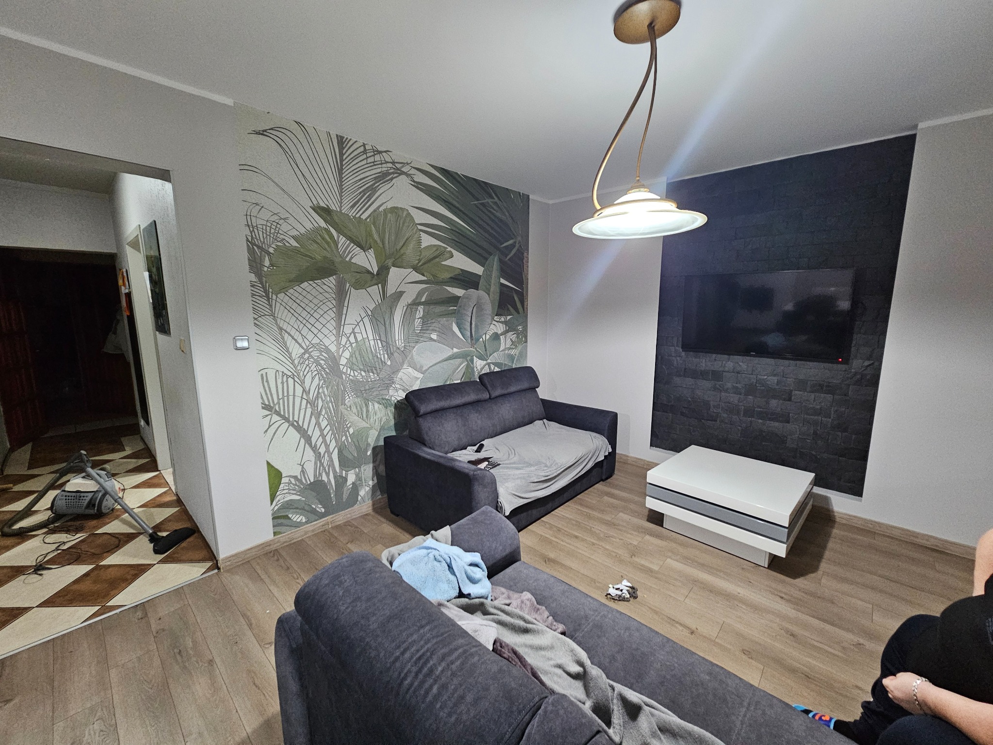 Salon z dwiema szarymi sofami, tapetą w tropikalne liście i czarną ścianą z telewizorem. Na podłodze panele, widoczny odkurzacz w przejściu do innego pomieszczenia.