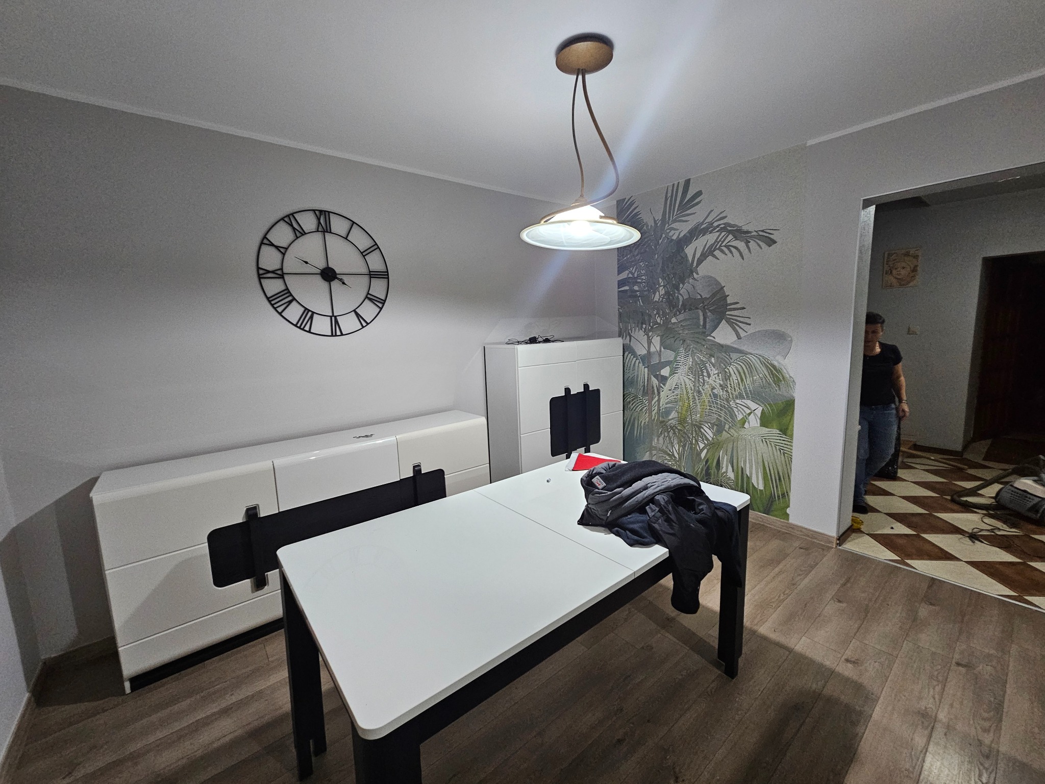 Wnętrze pokoju w Ostrowie Wielkopolskim z białymi meblami, zegarem na ścianie i dekoracyjną tapetą z motywem roślinnym. Na stole leżą ubrania.