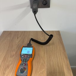 INNOTECH SP. Z O.O - Pomiar napięcia w gniazdku elektrycznym za pomocą miernika Sonel MPI-506 z czarnym przewodem spiralnym, widoczne wskazanie 250V na ekranie, na tle drewnianego blatu.
