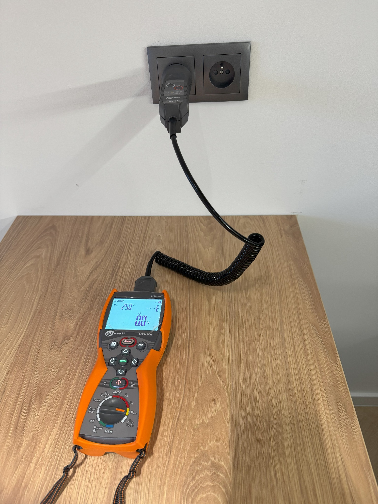 Pomiar napięcia w gniazdku elektrycznym za pomocą miernika Sonel MPI-506 z czarnym przewodem spiralnym, widoczne wskazanie 250V na ekranie, na tle drewnianego blatu.